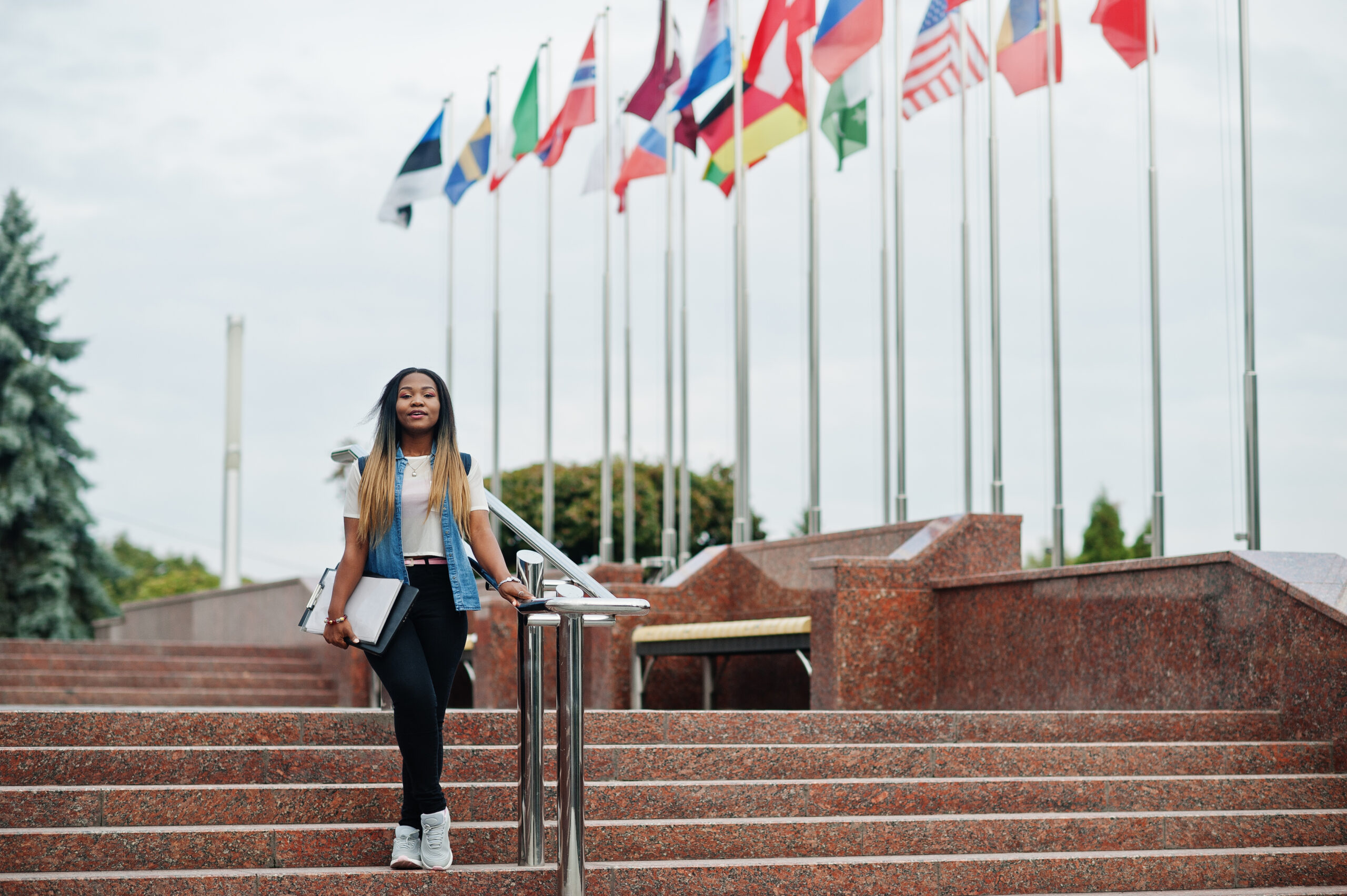 African student female posed with backpack and school items on yard of university, against flags of different countries.