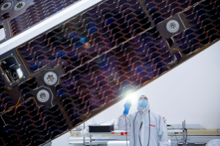 A technician in a white clean room gown and face mask shines a flashlight to inspect the dark, patterned cells of a large spacecraft solar array inside a clean room.