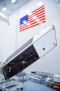 A large spacecraft sunshade and solar array panel rests angled upward on a test rig inside a high bay clean room, positioned beneath a large American flag on the wall.